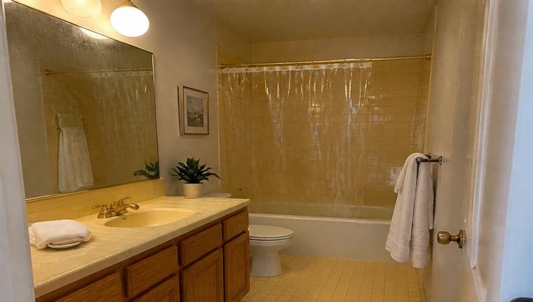 Bathroom with yellow tile, an oak vanity and a plastic tub curtain, before renovation.