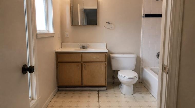 Bathroom with a small oak vanity, patterned vinyl floor and a dated medicine cabinet, before renovation.